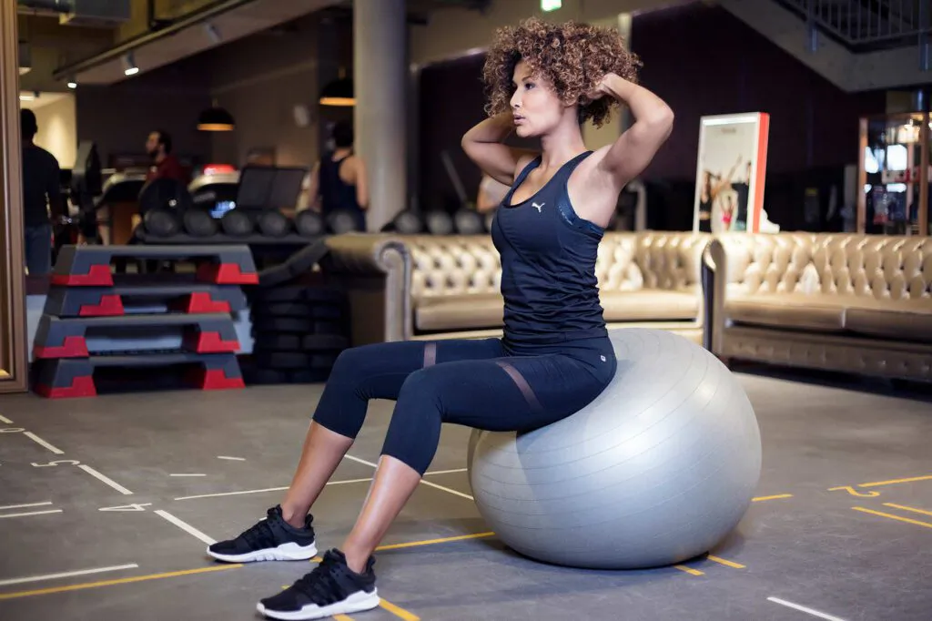 Marie Amière sitzt im Fitnessstudio auf einem silbernen Gymnastikball, die Hände am Hinterkopf.