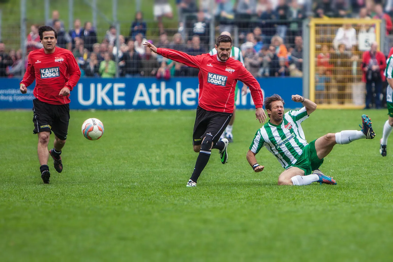 Zwei Fußballspieler in Rot laufen zum Ball, während ein Spieler in Grün-Weiß auf dem Rasen grätscht.