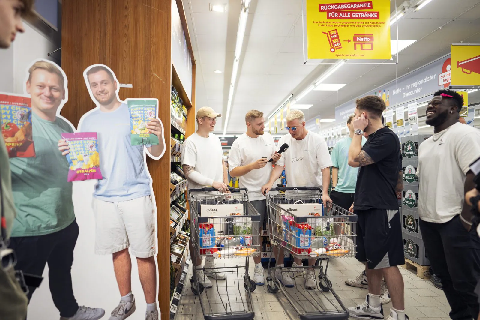 Männer mit Mikrofon und gefüllten Einkaufswagen im hellen Netto-Markt neben Pappaufstellern mit Happy-Chips-Tüten.