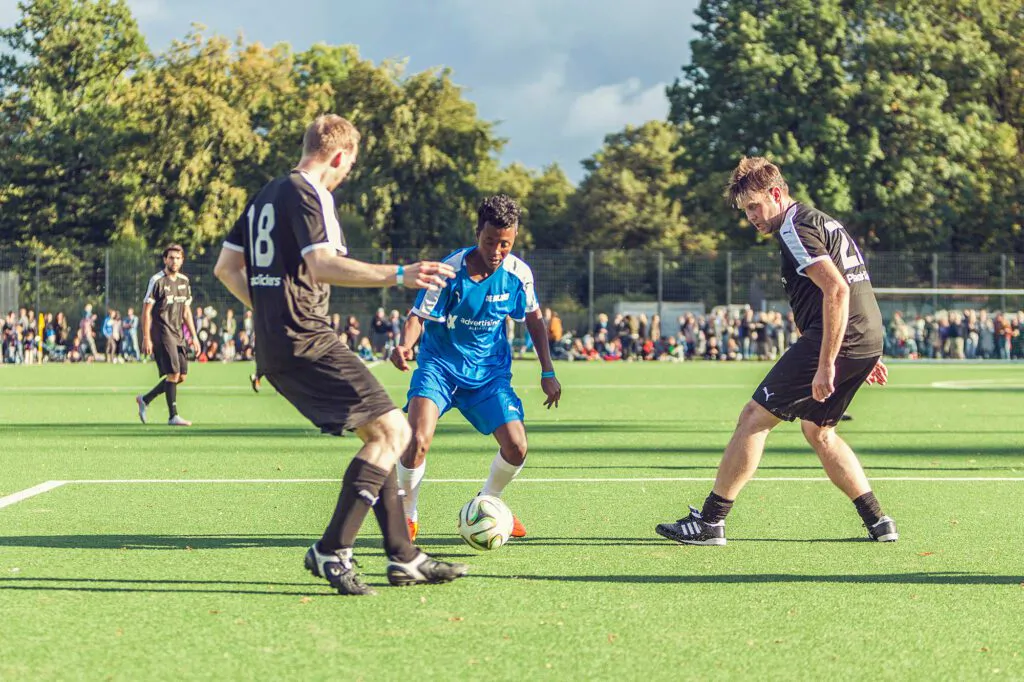Fußballer in Blau dribbelt zwischen zwei Gegnern in Schwarz auf sonnigem Kunstrasen, Zuschauer am Spielfeldrand.