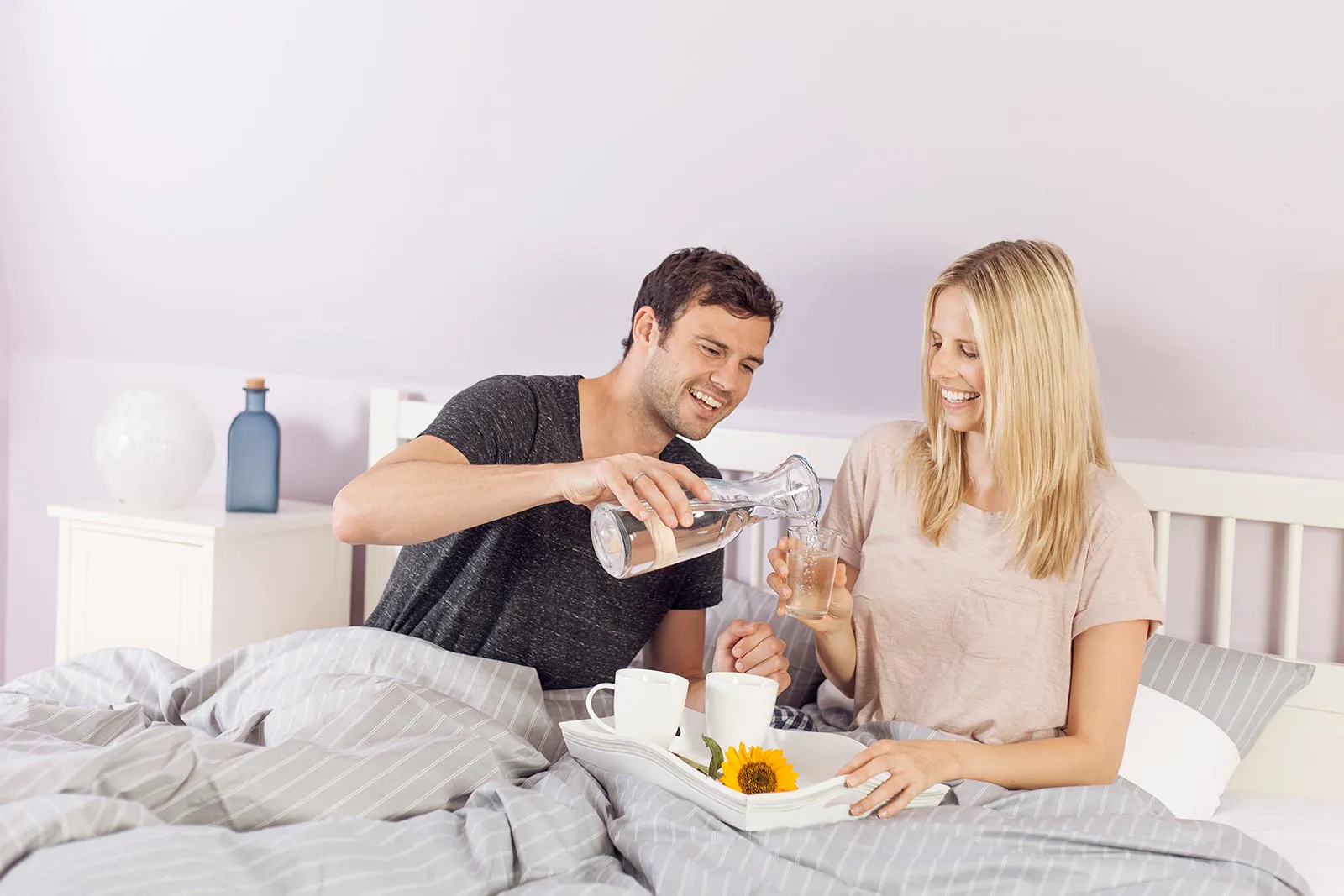 Mann schenkt einer lächelnden Frau im hellen Schlafzimmer Wasser ein; beide sitzen mit Frühstückstablett im Bett.