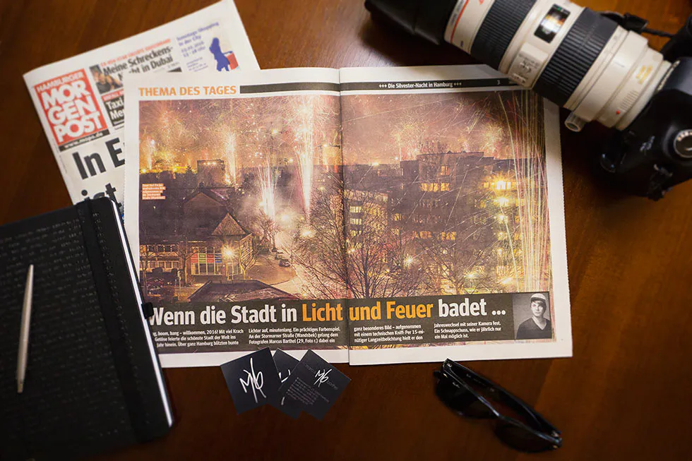 Aufgeschlagene Zeitung mit Feuerwerk über einer Stadt, daneben Kamera, Notizbuch und Brille auf einem Holztisch.