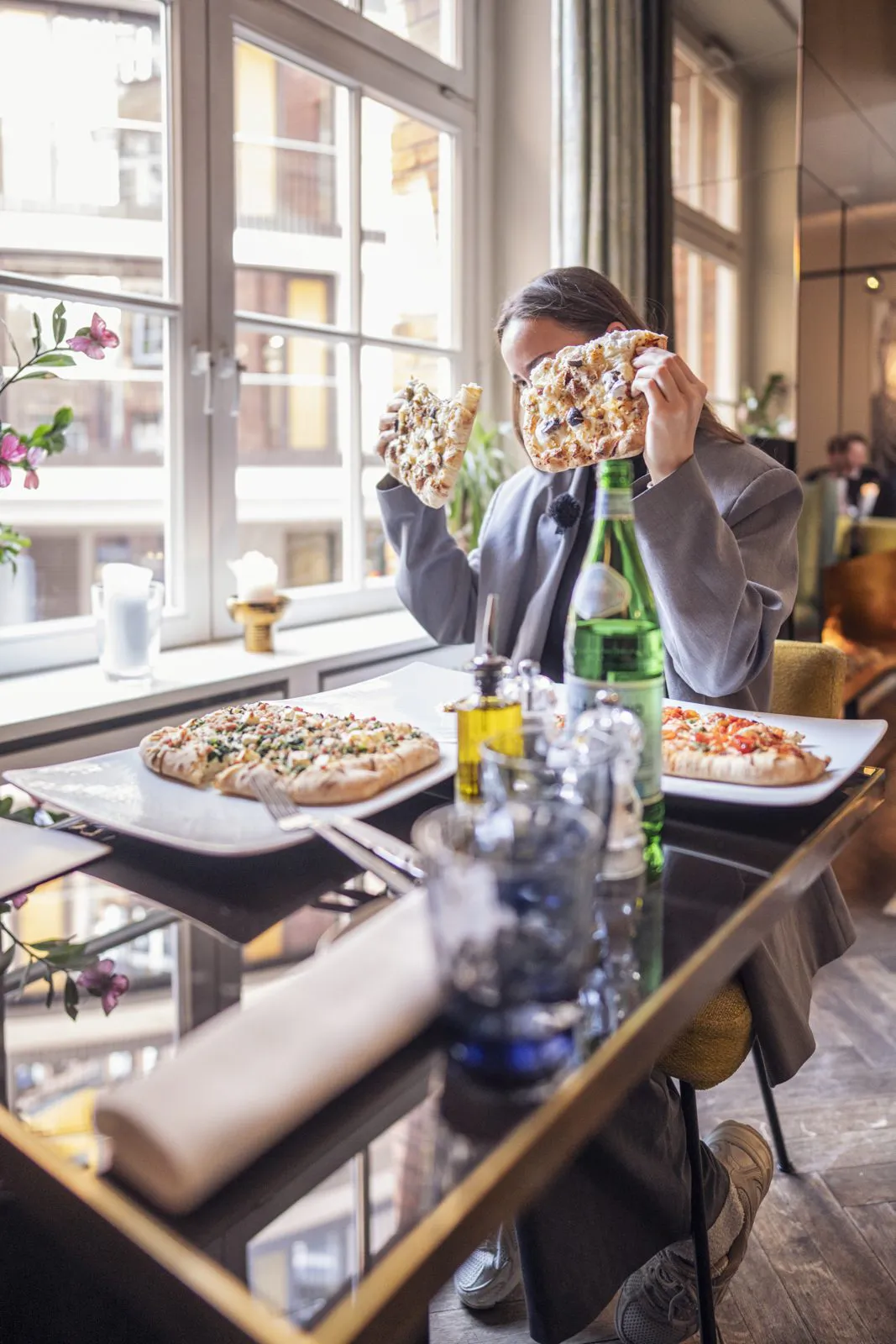 Person hält zwei Pizzastücke vor ihr Gesicht, auf dem Tisch liegen Pizzen im Tageslicht eines Restaurantfensters.