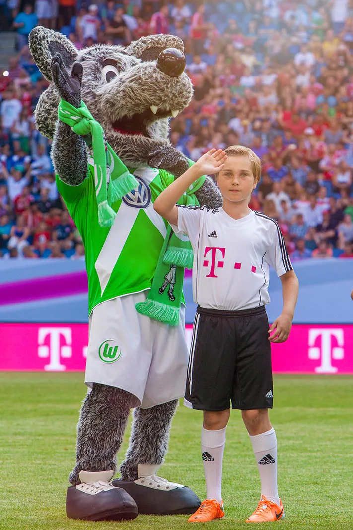 Junge im Fußballtrikot und Wolfsmaskottchen in Grün winken auf dem Rasen vor einer vollen Stadiontribüne.
