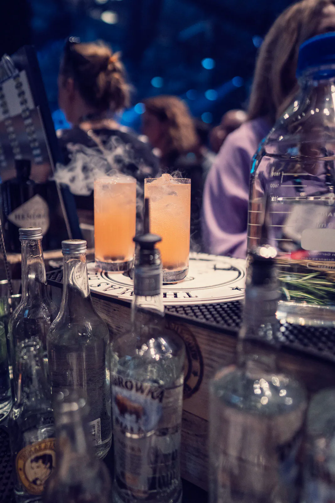 Zwei orange Cocktails mit Eis auf einer Bar, davor Spirituosenflaschen, dahinter Gäste im blauen Eventlicht.