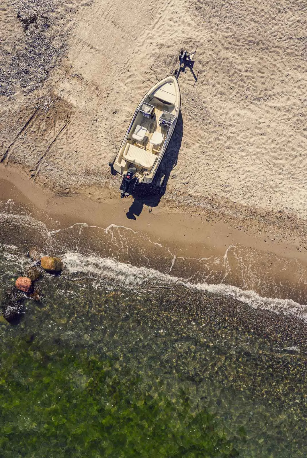 Luftaufnahme eines Motorboots am sonnigen Sandstrand mit kleinen Wellen und Steinen im klaren Wasser.