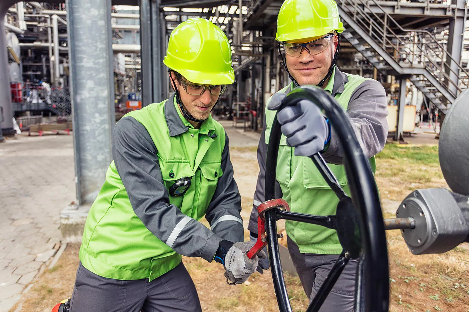 Zwei Personen mit Schutzhelmen und grüner Arbeitskleidung bedienen ein großes Handrad in der Raffinerie Heide.