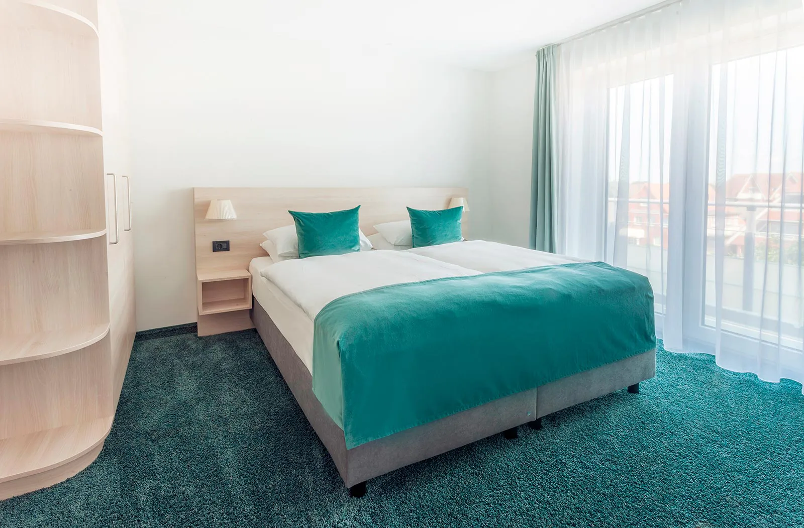 Helles Hotelzimmer mit Doppelbett, türkisfarbenen Kissen und Tagesdecke, Holzregal und bodentiefen Fenstern.