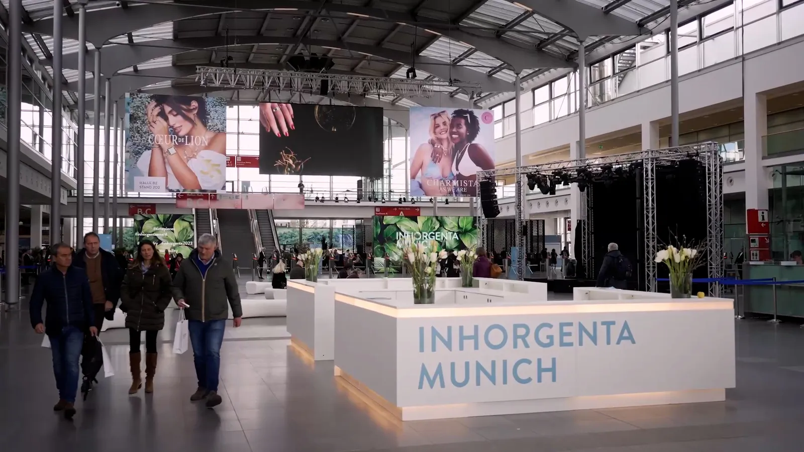Besucher gehen durch eine helle Messehalle mit weißen INHORGENTA-MUNICH-Theken, Blumen und großen Schmuckplakaten.