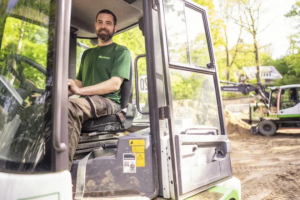 Lächelnder Mann im grünen Boymann-Shirt sitzt in der offenen Baggerkabine auf einer sonnigen Baustelle.