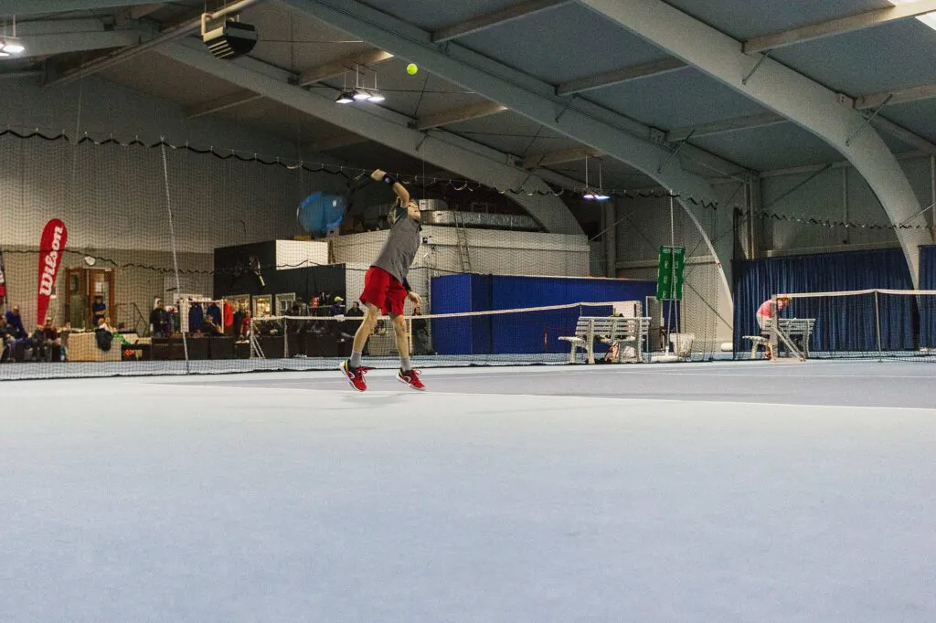 Tennisspieler in roter Hose springt beim Aufschlag unter künstlichem Licht in einer Tennishalle beim Wilson Master Cup.