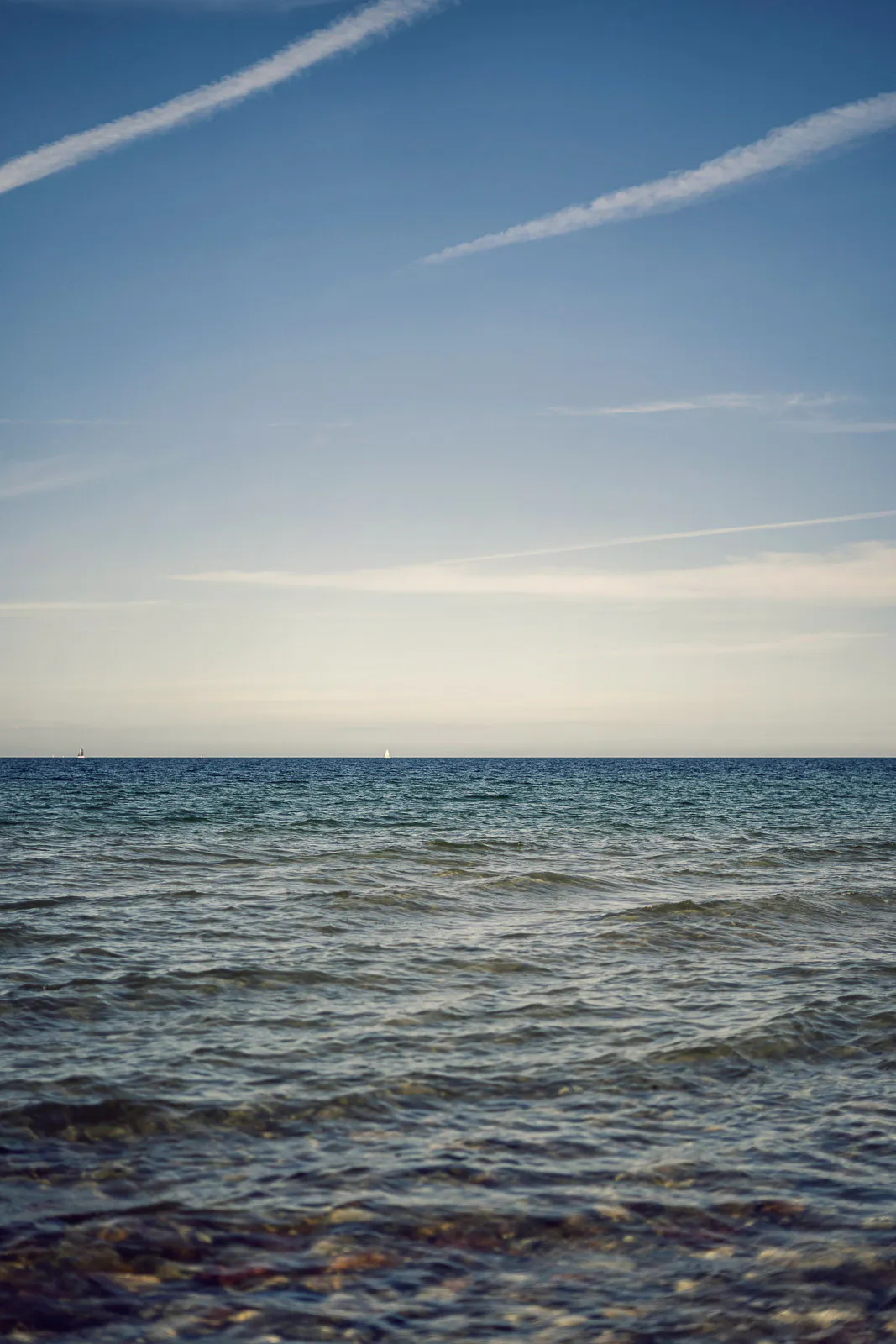 Kleine Wellen auf klarem Ostseewasser, Steine im Vordergrund und Kondensstreifen am blauen Himmel im sanften Licht.