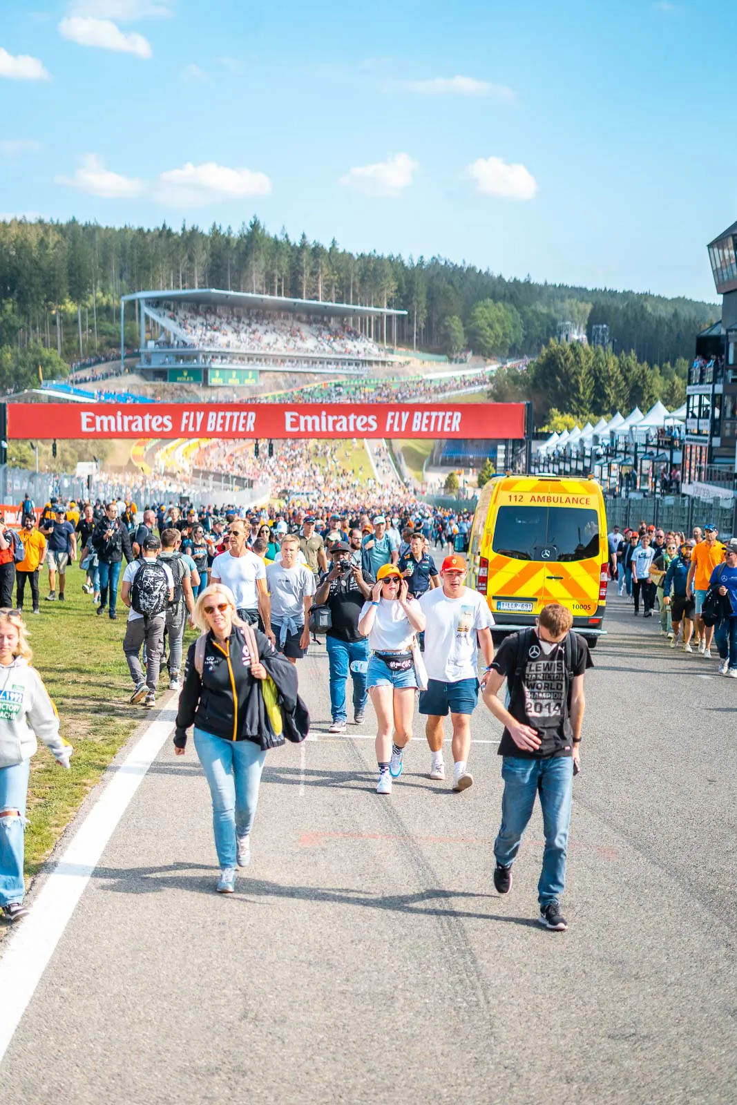 Zuschauer gehen bei Sonnenschein über eine Formel-1-Rennstrecke, neben ihnen ein Rettungswagen, dahinter Tribünen.