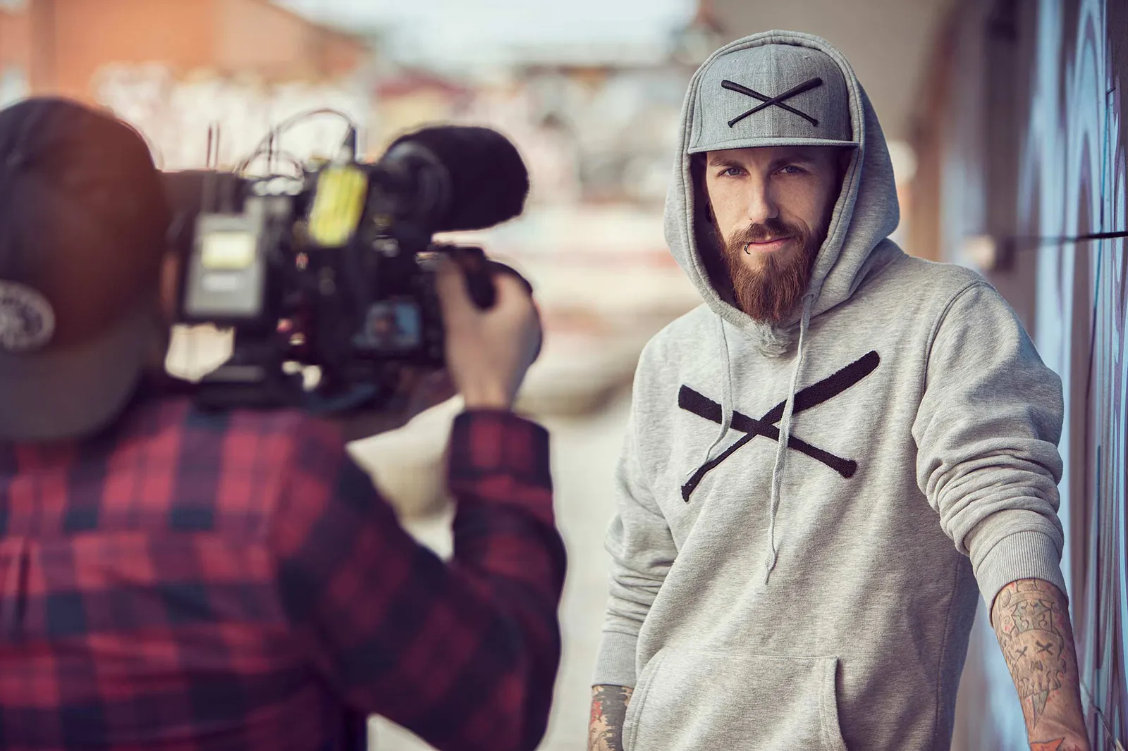 Dennis Grundt in grauem Hoodie und Kappe lehnt an einer Wand, während ein Kameramann ihn bei Tageslicht filmt.