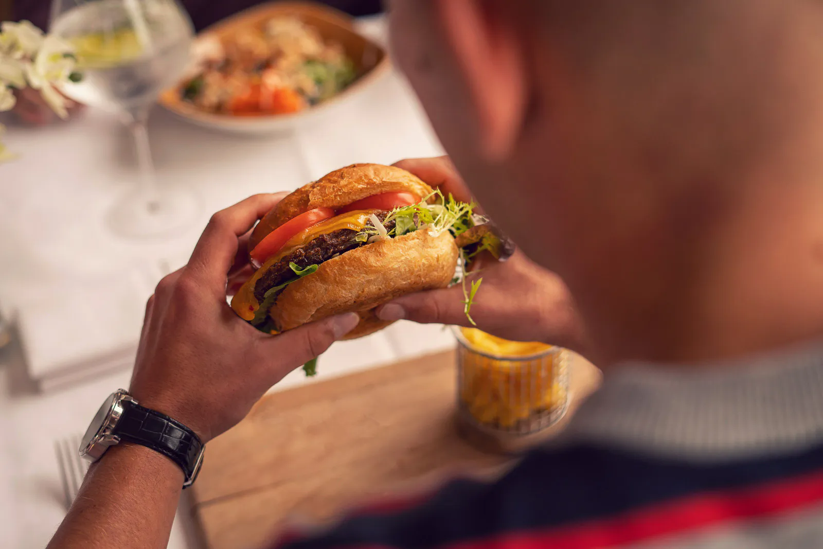 Gast hält einen Burger mit Käse, Tomate und Salat am gedeckten Tisch im Insel Beach Club Hannover.