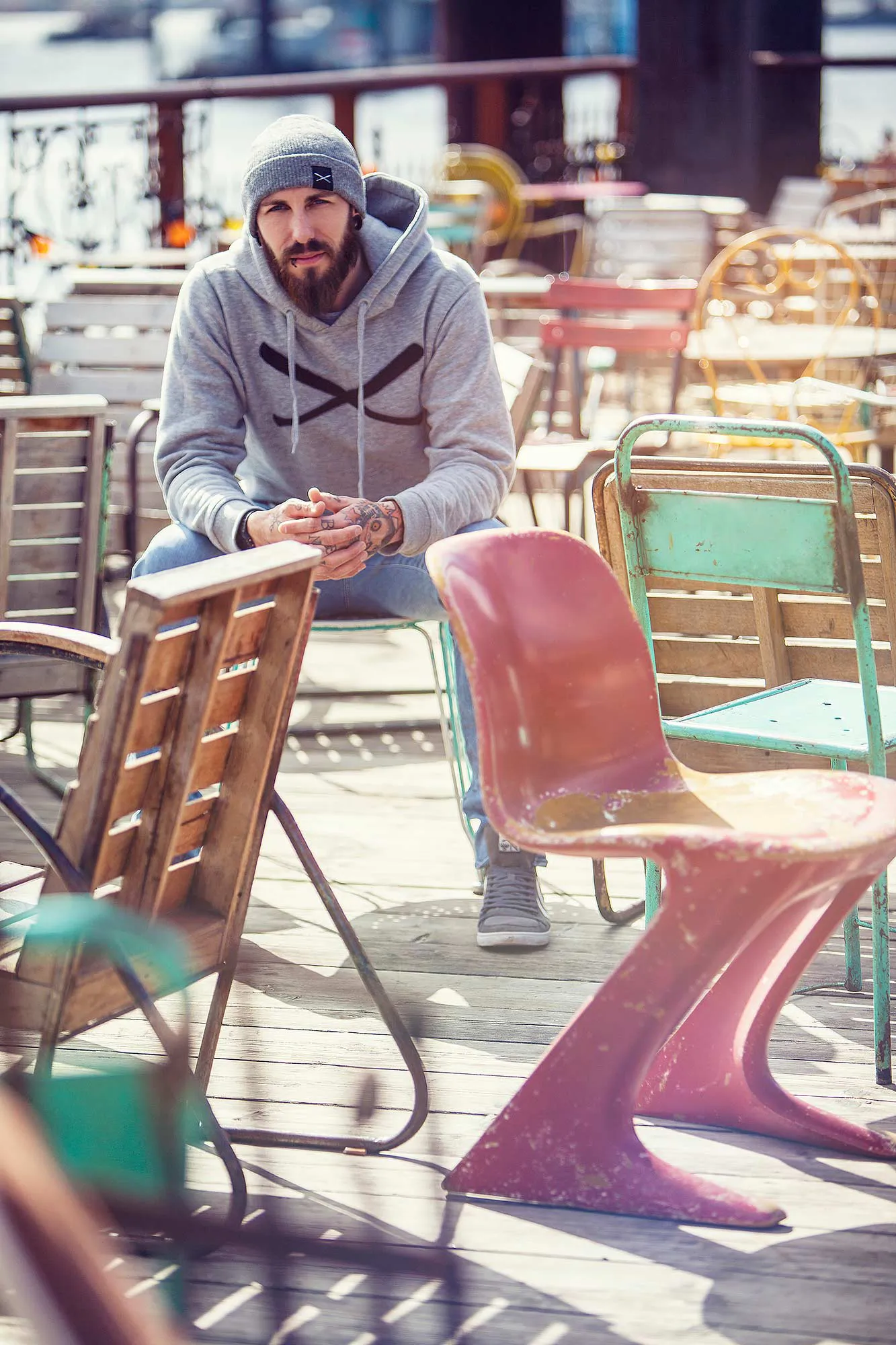 Bärtiger Mann mit Mütze und grauem Hoodie sitzt zwischen bunten Stühlen auf einer sonnigen Caféterrasse.