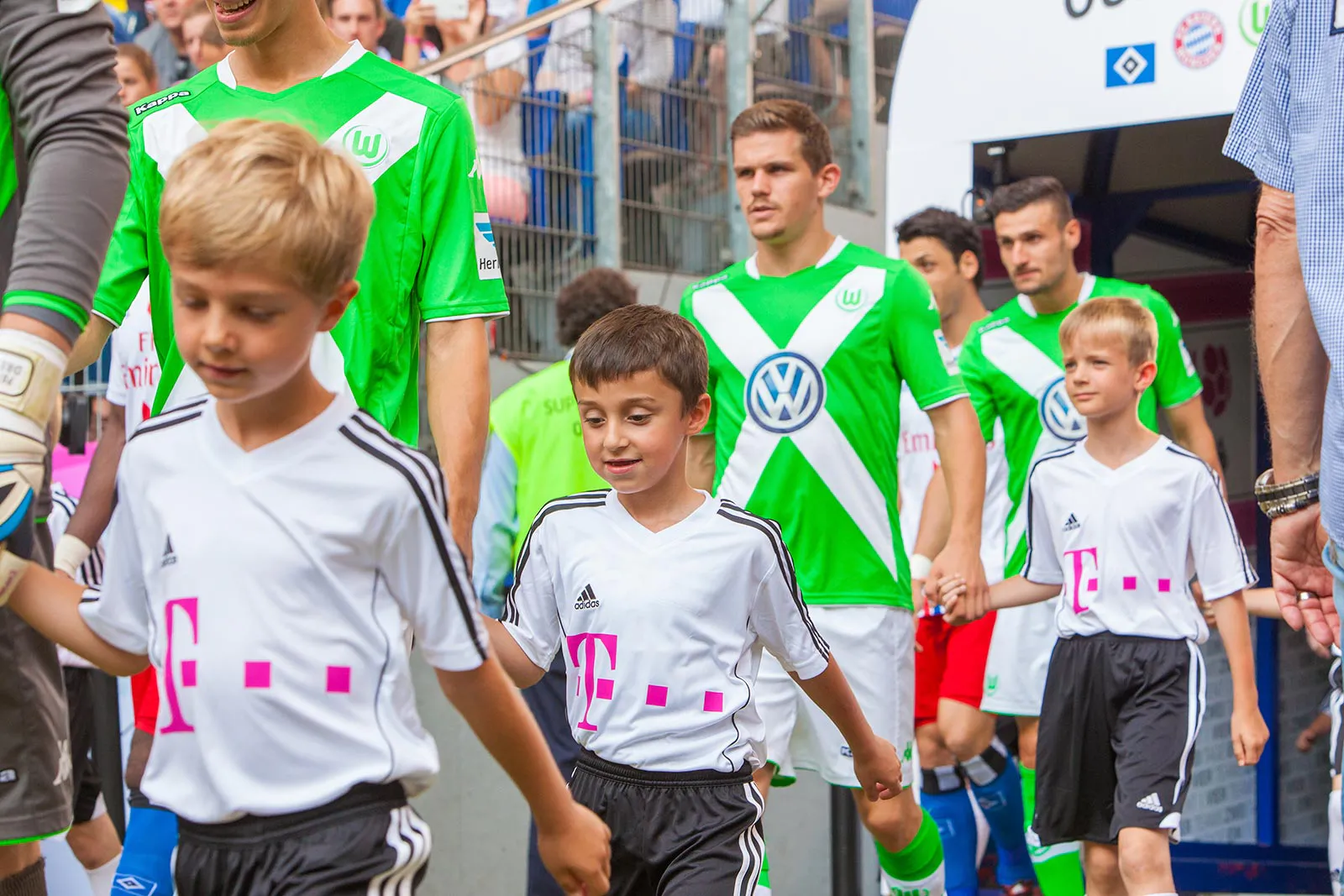 Fußballspieler in grünen Trikots laufen mit Kindern in weißen Trikots aus einem Stadiontunnel.