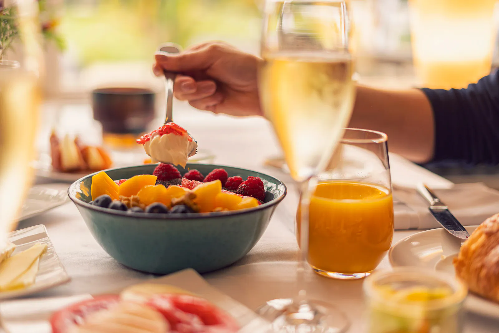 Hand löffelt Joghurt aus einer Schale mit Beeren und Mango; Orangensaft und Sekt stehen im warmen Licht auf dem Tisch.