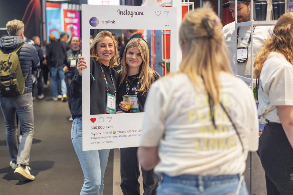Zwei Frauen posieren auf einer Messe mit einem Instagram-Fotorahmen, eine weitere Frau steht vor ihnen.