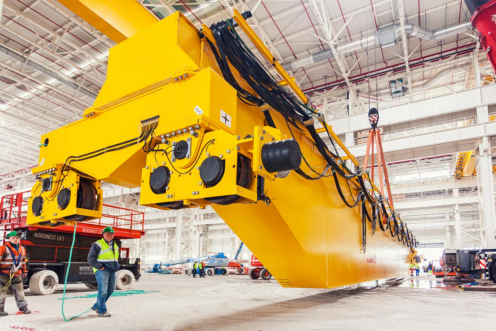 Arbeiter begleiten die Montage eines großen gelben KONECRANES-Kranträgers in einer hellen Industriehalle.