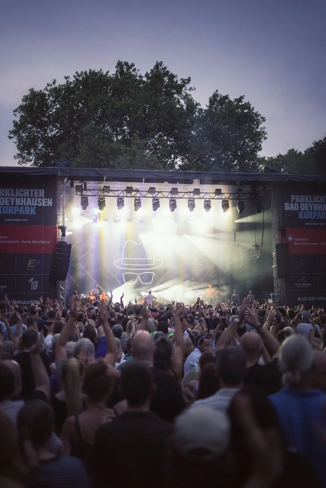 Publikum mit erhobenen Händen vor einer Open-Air-Bühne mit hellen Lichtstrahlen beim Konzert von Jan Delay.