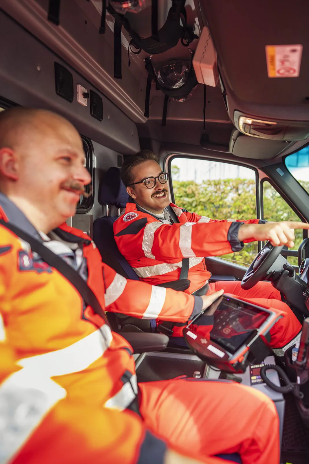 Zwei Johanniter-Rettungskräfte in orangefarbener Einsatzkleidung im Fahrzeug; eine zeigt nach vorn, eine hält ein Tablet.