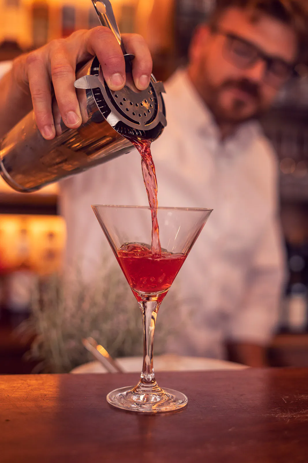 Barkeeper gießt einen roten Cocktail aus einem Shaker in ein Martiniglas an einer warm beleuchteten Bar.