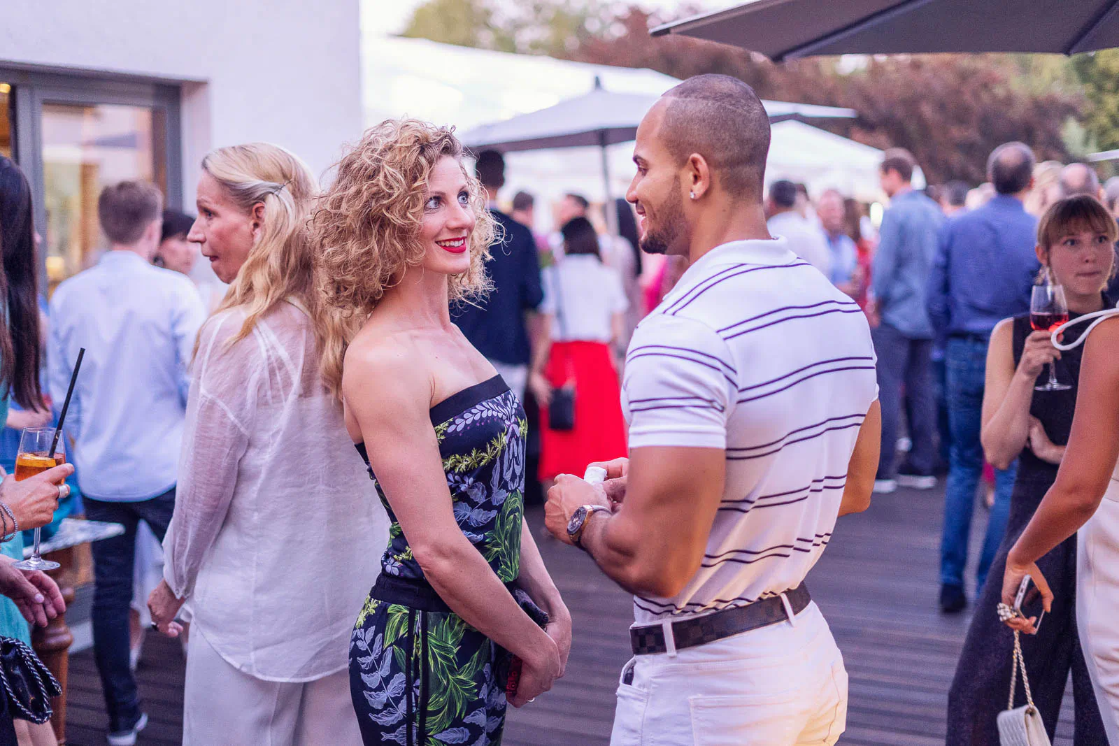 Frau im trägerlosen Kleid und Mann im gestreiften Poloshirt unterhalten sich bei einer Sommerparty auf einer Terrasse.