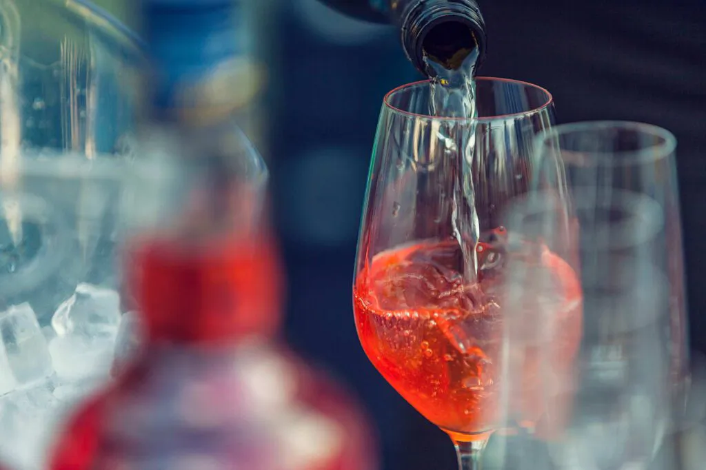 Eine klare Flüssigkeit wird aus einer Flasche in ein Weinglas mit orangefarbenem Drink und Eis gegossen.