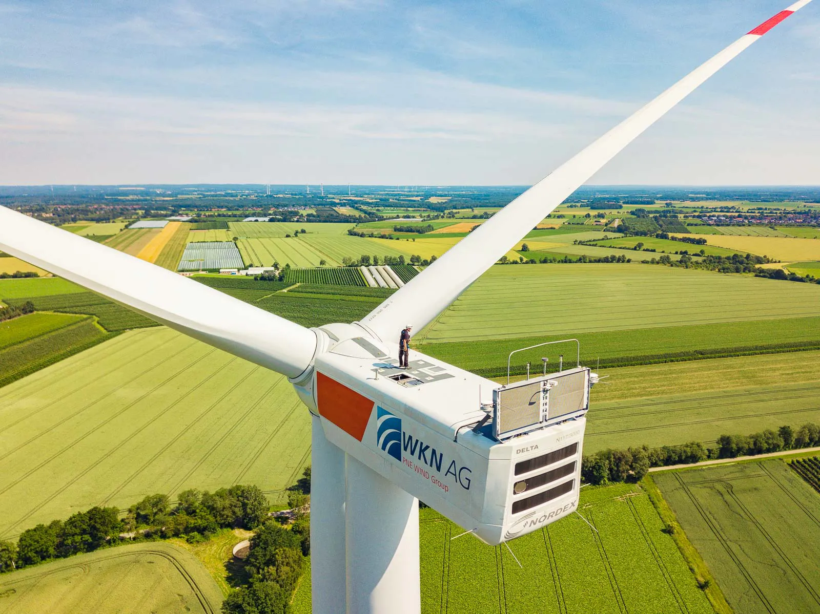 Person auf der Gondel einer Windkraftanlage hoch über grünen Feldern bei Tageslicht.