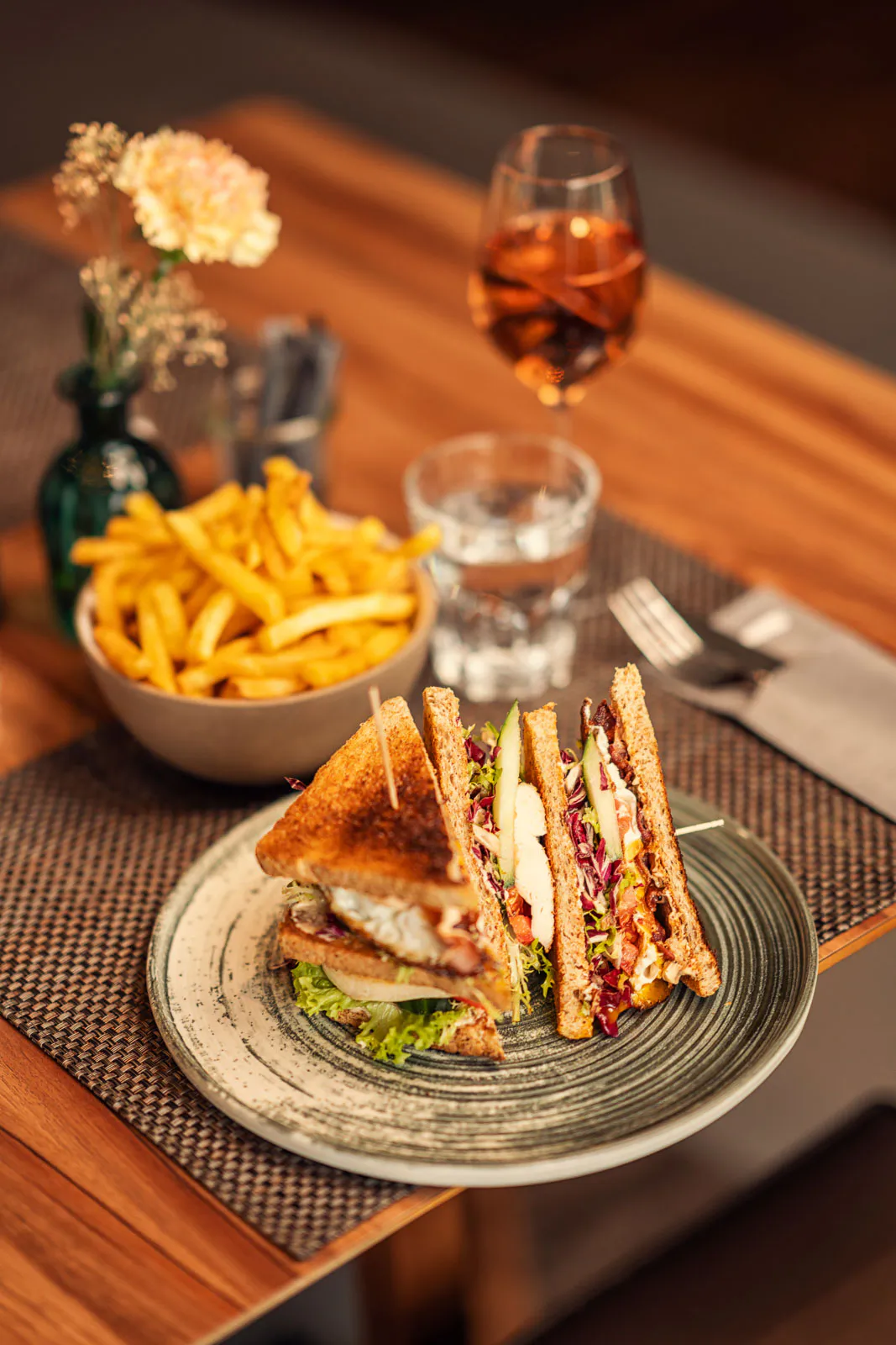Club-Sandwich auf einem Teller, daneben Pommes, Wasser und ein orangefarbenes Getränk auf einem warm beleuchteten Holztisch.