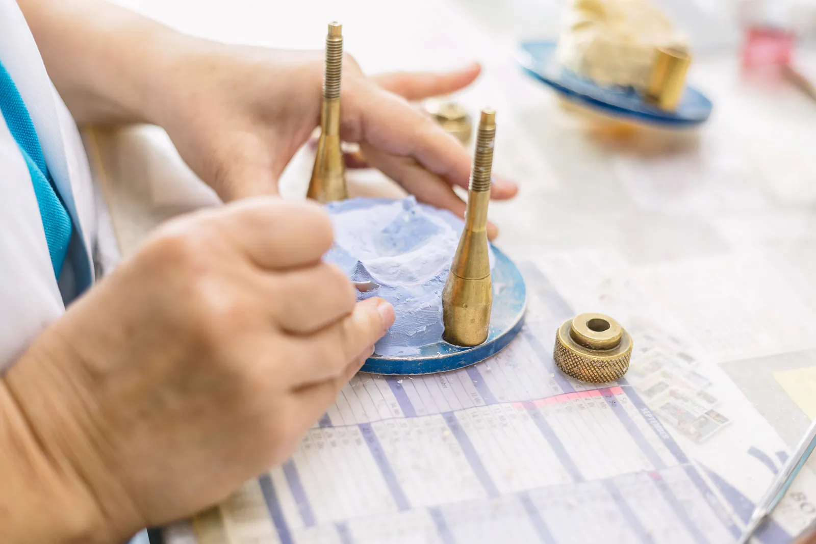 Hände bearbeiten ein blaues Gipsmodell in einer Metallhalterung am hell beleuchteten Arbeitsplatz im Dentallabor.