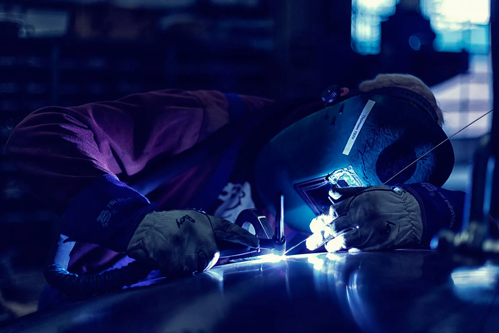Schweißer mit Schutzhelm und Handschuhen bearbeitet ein Metallteil im blauen Lichtbogen einer Werkhalle.