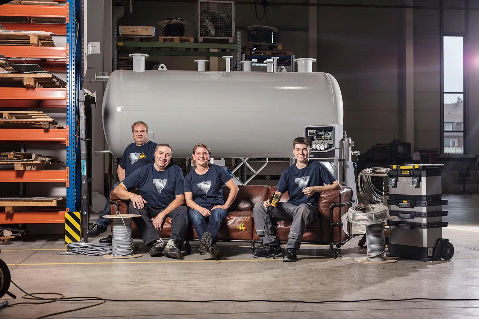 Vier Männer in Arbeitskleidung an einem braunen Ledersofa in einer Werkhalle vor einem großen Metalltank.