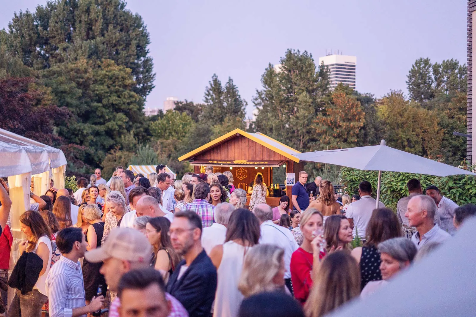 Gäste unterhalten sich bei einer Sommerparty im Freien vor einer Holzhütte, umgeben von Bäumen und Sonnenschirmen.
