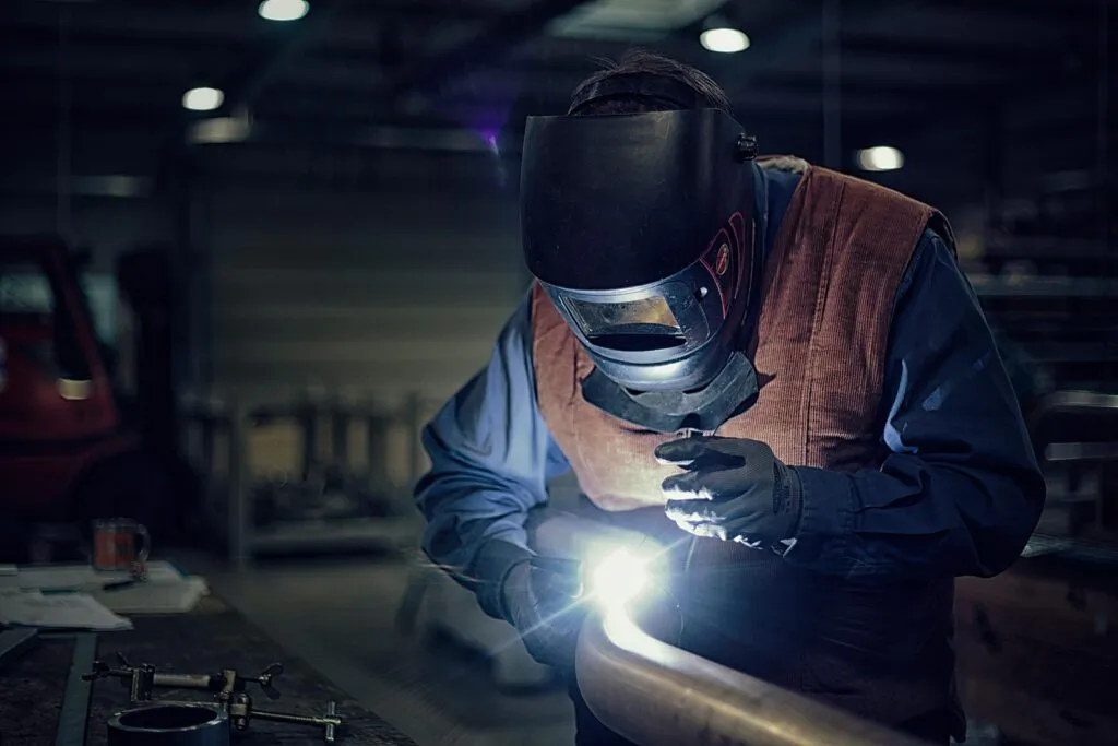 Schweißer mit Schutzhelm und Lederweste bearbeitet ein Metallrohr im hellen Lichtbogen einer dunklen Werkhalle.