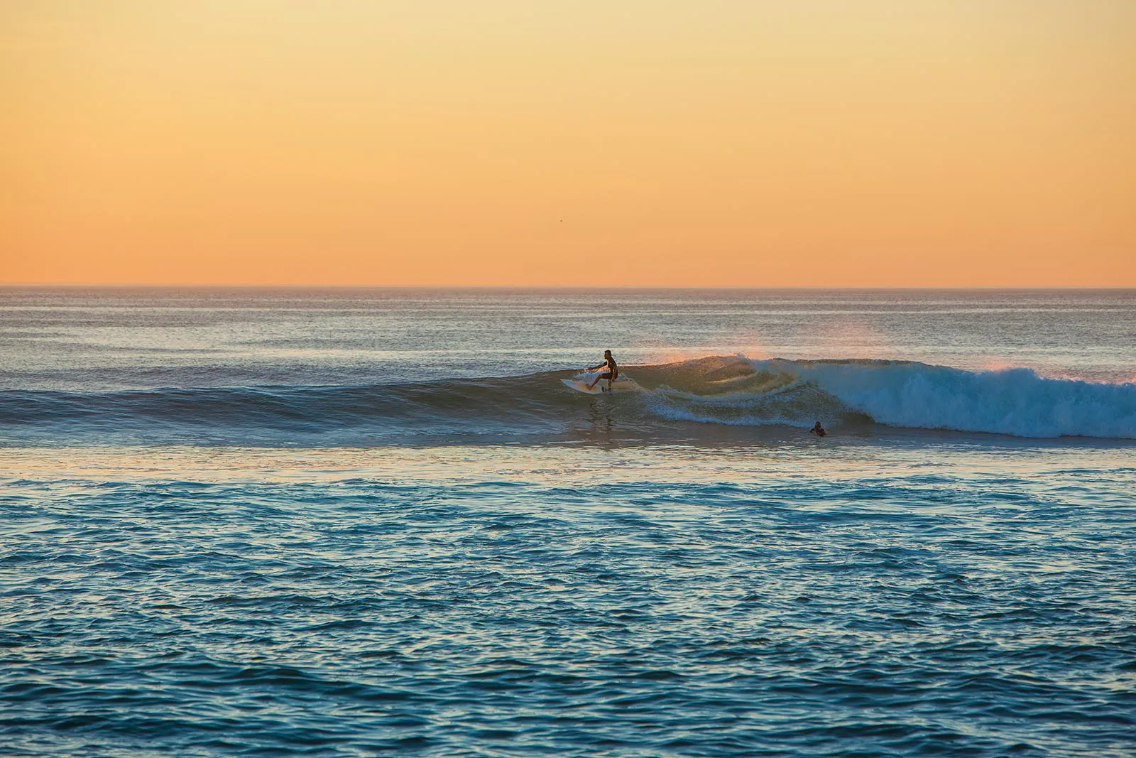 Eine Person surft auf einer brechenden Welle im Meer, eine weitere wartet im Wasser unter orangefarbenem Abendhimmel.