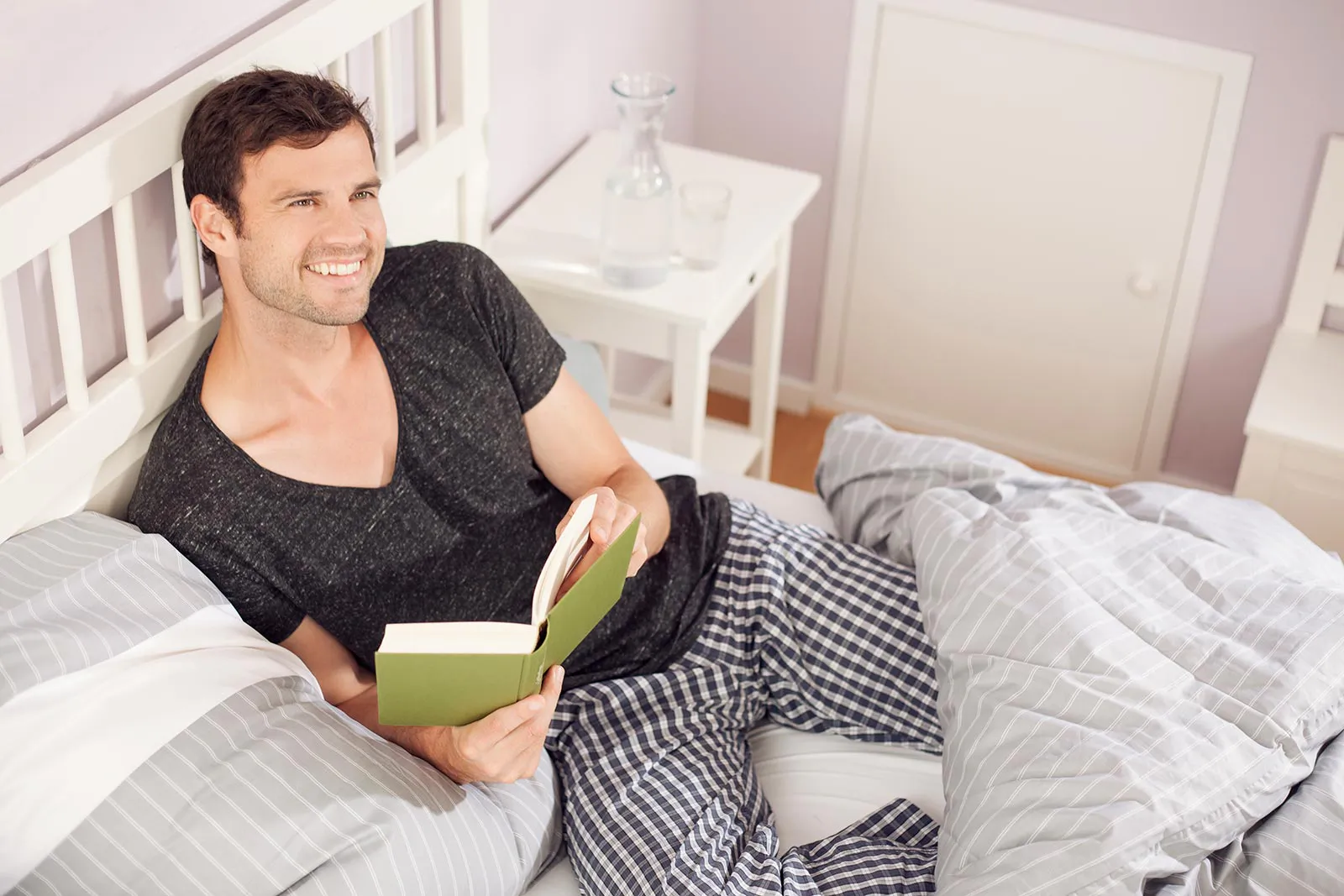 Lächelnder Mann mit offenem Buch im Bett, umgeben von grau-weiß gestreifter Bettwäsche im hellen Schlafzimmer.