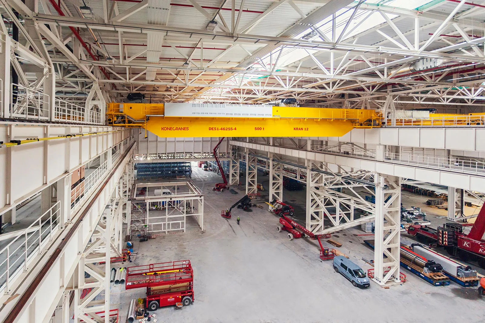 Gelber KONECRANES-Brückenkran in heller Industriehalle mit weißen Stahlträgern und roten Hebebühnen.