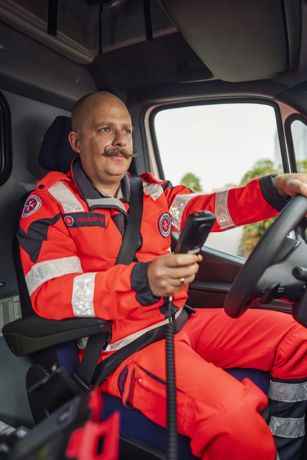 Johanniter-Mitarbeiter in roter Einsatzkleidung sitzt am Steuer eines Einsatzfahrzeugs und hält ein Funkgerät.