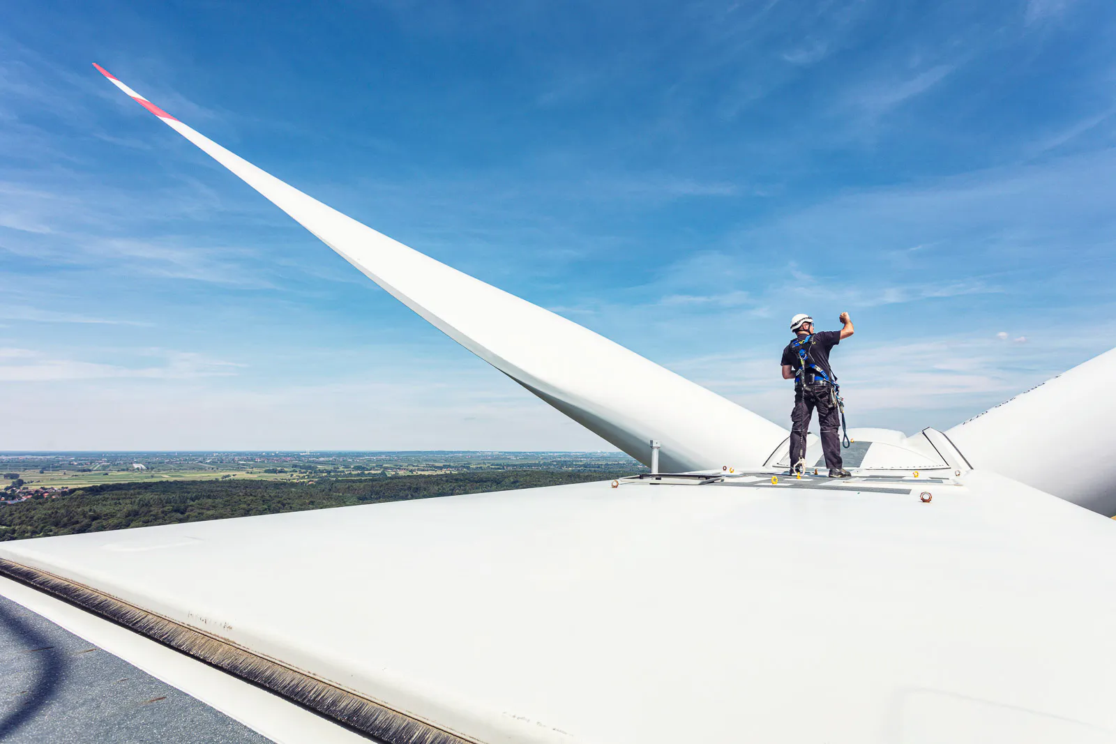 Person mit Helm und Sicherungsgurt auf einer Windkraftanlage neben einem Rotorblatt unter blauem Himmel.