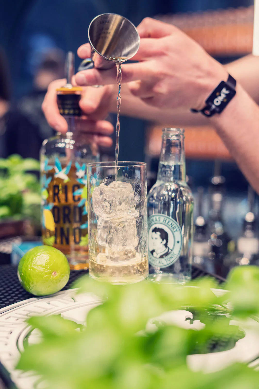 Hände gießen an einer Bar Flüssigkeit ins Glas mit Eis; daneben stehen eine Thomas-Henry-Flasche und eine Limette.