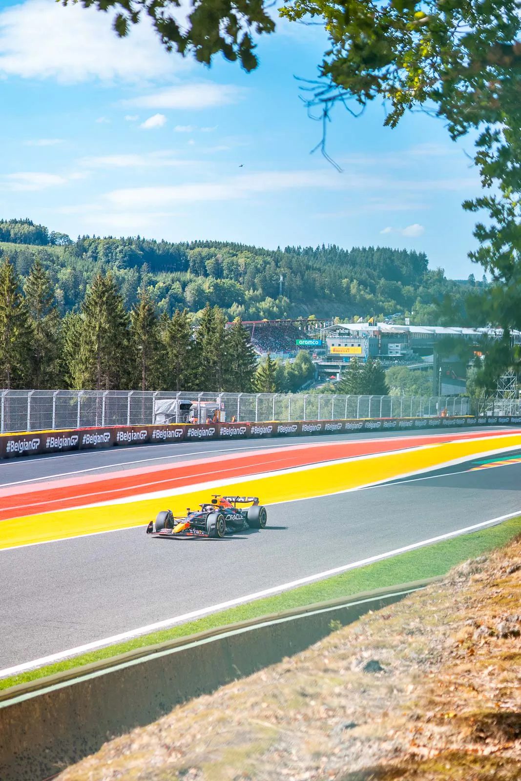 RedBull-Racing-F1-Wagen auf sonniger Rennstrecke mit rot-gelben Randstreifen, umgeben von bewaldeten Hügeln.
