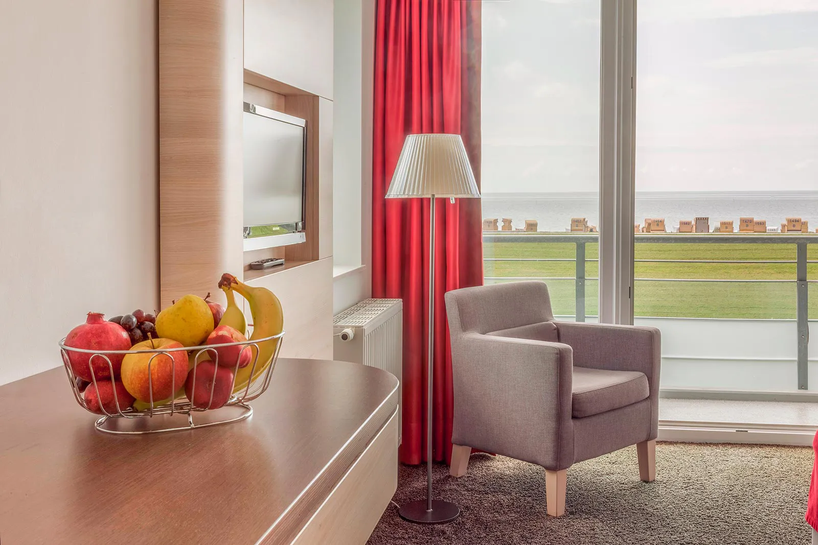 Helles Hotelzimmer mit Obstkorb, Sessel und Stehlampe vor roten Vorhängen, Balkon mit Blick ins Weite.