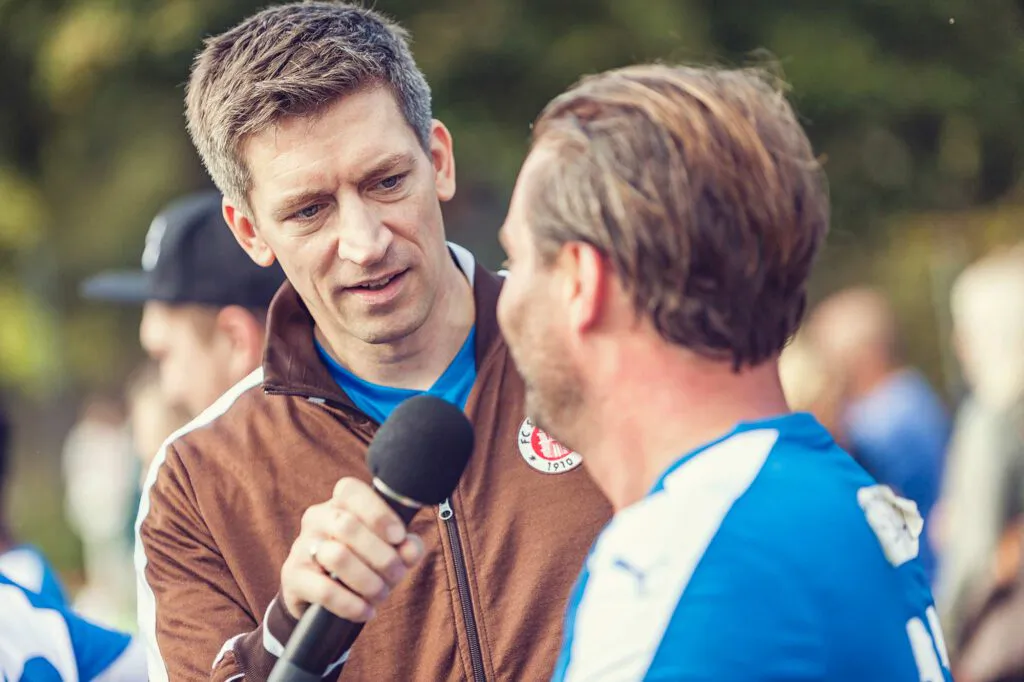 Mann mit Mikrofon interviewt einen Mann im blau-weißen Fußballtrikot im Freien bei Tageslicht.