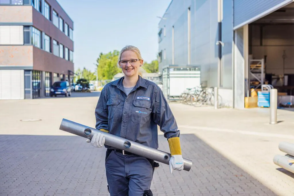 Frau in Arbeitskleidung und Schutzhandschuhen trägt ein Metallrohr vor einer sonnenbeschienenen Industriehalle.