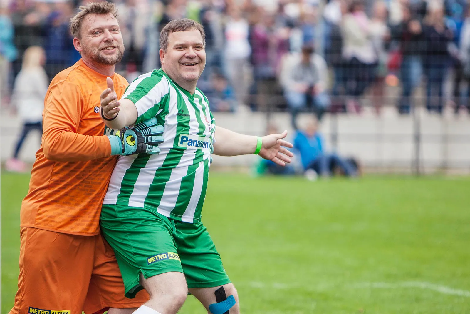 Zwei lachende Fußballspieler in Orange und Grün-Weiß umarmen sich auf dem Rasen vor Publikum.