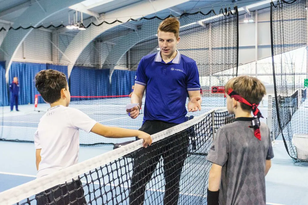 Ein Mann im blauen Poloshirt spricht mit zwei Jungen am Tennisnetz in einer hell beleuchteten Halle.