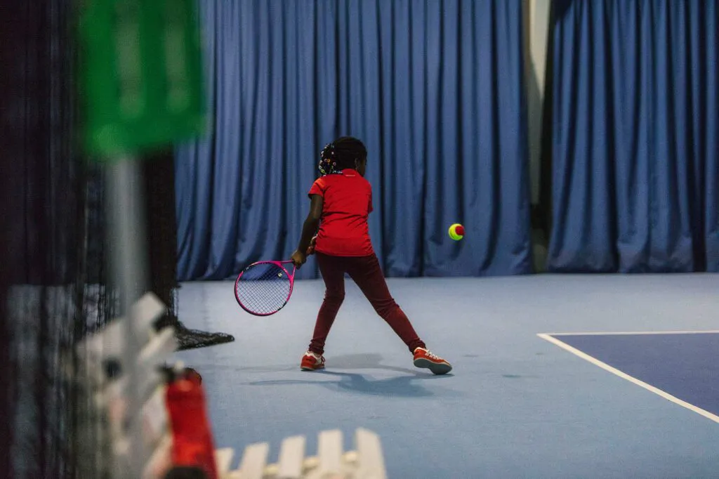 Kind in rotem Shirt mit Tennisschläger vor einem Ball auf blauem Hallenplatz beim Wilson Master Cup.
