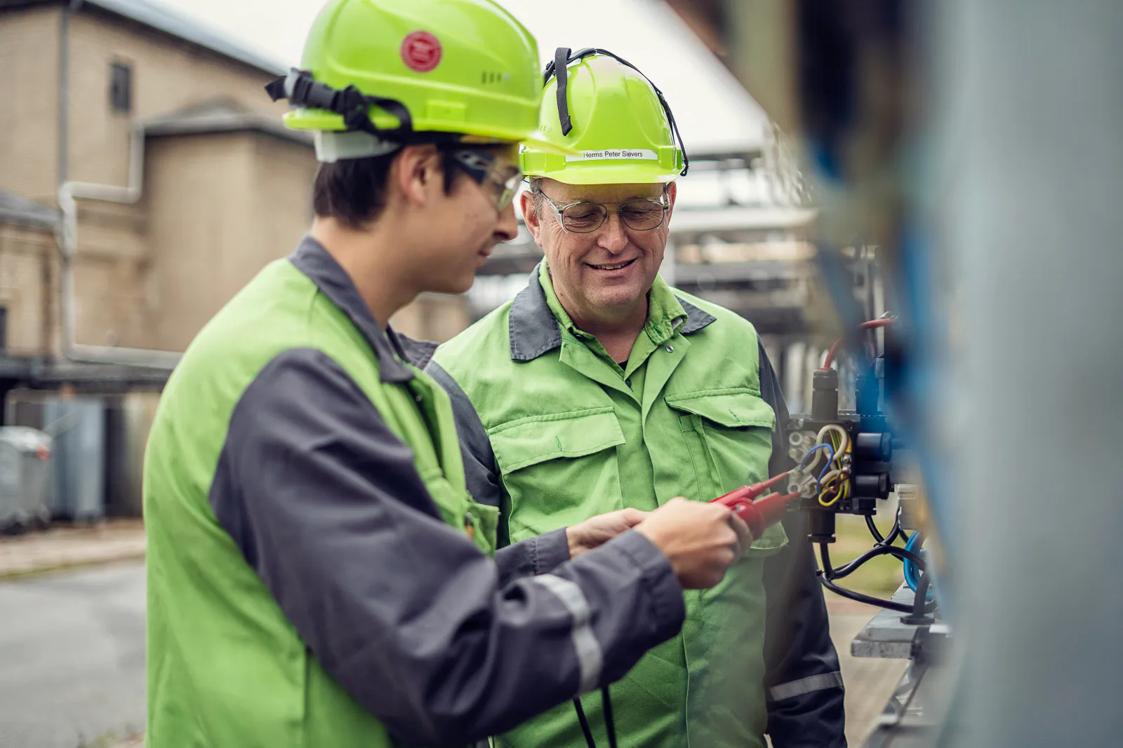 Zwei Männer in grüner Arbeitskleidung und Schutzhelmen arbeiten bei Tageslicht an einer Anlage der Raffinerie Heide.